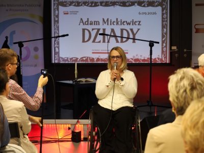 Kobieta w okularach i jasnej bluzce przemawia do mikrofonu, siedząc na wózku inwalidzkim na scenie. W tle znajduje się ekran z napisem „Narodowe Czytanie. Adam Mickiewicz DZIADY. 6.09.2026” oraz roll-up biblioteki. Z przodu widoczny jest mężczyzna nagrywający wystąpienie smartfonem oraz publiczność