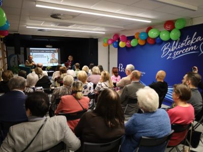 Wypełniona po brzegi sala wykładowa z niebieską ścianą ozdobioną kolorowymi balonami i napisem „Biblioteka z sercem”. Z przodu na fotelach siedzą prelegenci, a w tle widać ekran z prezentacją. Widownia obracająca się tyłem do obiektywu słucha wystąpienia