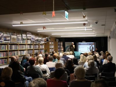 Szerokie ujęcie sali bibliotecznej wypełnionej publicznością podczas spotkania autorskiego. Na końcu sali, na podwyższeniu, stoi Jacek Fedorowicz z mikrofonem w ręku. W tle znajduje się ekran z napisem „Spotkanie autorskie z Jackiem Fedorowiczem”. Wzdłuż lewej ściany ciągną się wysokie regały z książkami