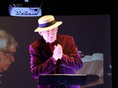 Zbliżenie na Stanisława Górkę stojącego przy pulpicie nutowym. Artysta ma na sobie słomkowy kapelusz z czarną wstążką oraz purpurową marynarkę, składa dłonie do oklasków i patrzy w stronę publiczności. W tle widać fragment ekranu projekcyjnego