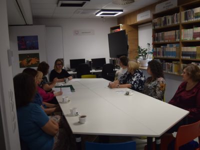 Kobiety uczestniczące w spotkaniu siedzą wokół długiego, białego stołu w bibliotece. Perspektywa ujęcia ukazuje wnętrze sali z widocznymi w tle stanowiskami komputerowymi oraz regałami z książkami. Na stole stoją filiżanki z napojami oraz dokumenty w zielonym segregatorze