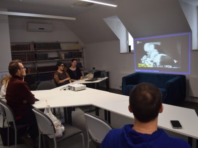 Spotkanie kawiarenki językowej w oddziale dla dorosłych. Wolontariuszki i uczestnicy siedzą przy białych stołach w kształcie litery V, otoczeni regałami z książkami. Na ścianie wyświetlany jest czarno-biały film z włoskim napisem „10 DICEMBRE 1926”. Dwie wolontariuszki siedzące przy laptopie moderują pokaz