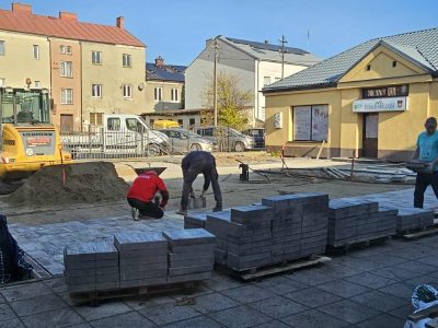 Robotnicy układający szarą kostkę brukową na wyrównanym podłożu placu przed „Domem Harcerza”. Na pierwszym planie ustawiono rzędy drewnianych palet szczelnie załadowanych kostką brukową. Dwaj pracownicy w ciemnych ubraniach (jeden w czerwonej bluzie) klęczą na ułożonym już fragmencie nawierzchni, dopasowując kolejne elementy. Po lewej stronie placu stoi żółta ładowarka kołowa marki Liebherr