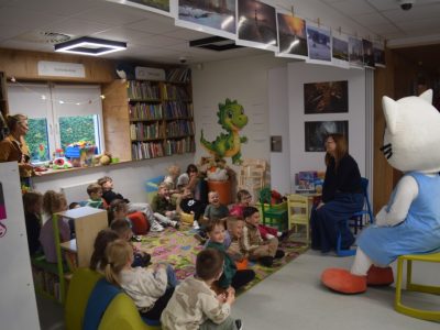 Bibliotekarka w okularach siedzi na niskim krześle i rozmawia z grupą dzieci siedzących na kolorowym dywanie w strefie dla najmłodszych. Obok niej na żółtym krześle siedzi duża maskotka Kici Koci. Dzieci słuchają z zaciekawieniem, a w tle widać regały z zabawkami i książkami oraz wystawę zdjęć