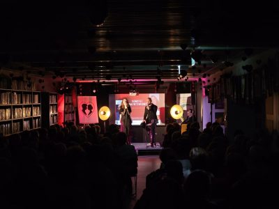 Szeroki kadr na zaciemnioną salę koncertową Miejskiej Biblioteki Publicznej w Przasnyszu wypełnioną publicznością. Pod sufitem wiszą dekoracyjne papierowe serduszka. Na oświetlonej różowym i ciepłym światłem scenie występują Natalia Srokocz i Jerzy Grzechnik