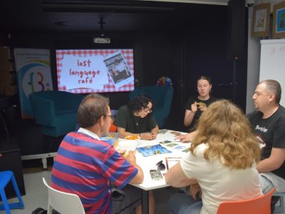Pięcioro uczestników siedzi wokół białego stołu, grając w planszową grę językową. Na ekranie projektora w tle wyświetla się plansza z napisem "last language café". Na stole, oprócz elementów gry, leżą telefony komórkowe oraz miseczki z przekąskami. Uczestnicy są skupieni, rozmawiają i wskazują karty na planszy