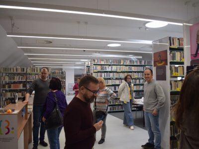 Wnętrze biblioteki z wysokimi regałami książek. Przy ladzie recepcyjnej z logo biblioteki stoi mężczyzna rozmawiający z kilkoma osobami. W przestrzeni między regałami widoczni są inni uczestnicy spotkania oraz plakaty z fotografiami na ścianach