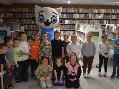 Pamiątkowe zdjęcie grupy dzieci z maskotką Kici Koci w bibliotece. Wielka maskotka w niebieskiej sukience stoi w centrum, otoczona uśmiechniętymi dziećmi. Część dzieci stoi, a kilka dziewczynek klęczy w pierwszym rzędzie na tle regałów wypełnionych książkami