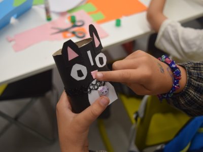 Zbliżenie na dłonie dziecka trzymającego figurkę czarnego kota zrobioną z tekturowej rolki. Zabawka ma naklejone papierowe uszy, biało-czarne oczy, różowy nosek oraz założony czarny naszyjnik z małą zawieszką