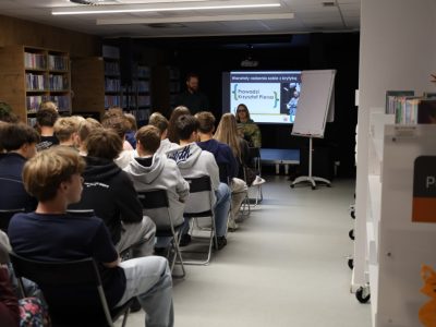 Widok z tyłu na grupę młodzieży siedzącą w sali bibliotecznej. Przed widownią, przy ekranie z prezentacją Krzysztofa Piersy, siedzi dyrektor biblioteki przemawiająca do uczestników