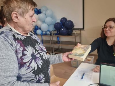 Starsza kobieta w szarym wzorzystym swetrze przekazuje książkę kobiecie w okularach siedzącej przy biurku z laptopem. W tle widoczne niebieskie i białe balony