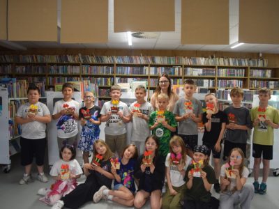 Wspólne zdjęcie uczestników warsztatów w bibliotece. Piętnaścioro dzieci oraz stojąca w środku dorosła kobieta w okularach pozują na tle regałów z książkami. Każde dziecko trzyma w rękach własnoręcznie zrobioną z kolorowego kartonu laurkę przedstawiającą tulipany w doniczce