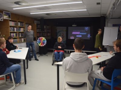 Wieczór quizowy w bibliotece. Bibliotekarz stoi obok kolorowego koła fortuny i patrzy na uczestników siedzących przy białych stołach. W tle dwie wolontariuszki przy flipcharcie oraz ekran z napisem „Wieczór Quizowy”. Na środku sali, tyłem do obiektywu, siedzi kobieta na wózku