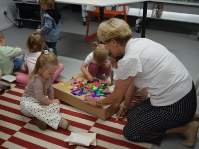 Warsztaty sensoryczne dla najmłodszych. Grupa małych dzieci wraz z opiekunką klęczą na pasiastym dywanie wokół kartonowego pudełka wypełnionego mnóstwem drobnych, kolorowych piankowych elementów