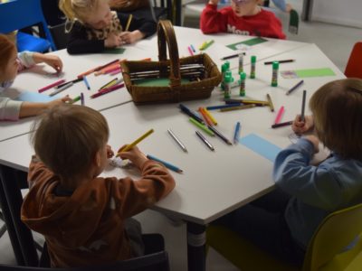 Zbliżenie na grupę dzieci pracujących przy wspólnym stole. Na środku stoi wiklinowy koszyk, wokół którego leżą liczne kredki i kleje. Dzieci starannie kolorują i ozdabiają papierowe zakładki do książek
