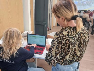 Ujęcie od tyłu. Kobieta z blond kręconymi włosami w ciemnej bluzie z napisem „BIBLIOTEKA” obsługuje czerwony laptop. Obok niej stoi druga kobieta w bluzce w zebrany wzór paisley i przygląda się ekranowi. W tle widać pozostałych uczestników spotkania siedzących w sali