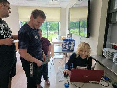 Kobieta o blond włosach siedzi przy stanowisku komputerowym i obsługuje myszką czerwonego laptopa. Obok niej stoją trzej uczestnicy spotkania, w tym mężczyzna w ciemnej koszulce, który z uśmiechem przygląda się pracy na komputerze