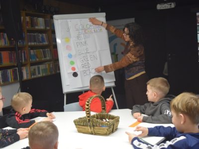 Grupa chłopców siedzi przy białym stole, słuchając wolontariuszki. Kobieta stoi przy tablicy flipchart, na której widnieje napis „COLORS IN ENGLISH” oraz lista nazw kolorów z odpowiadającymi im kolorowymi kropkami. Na stole stoi wiklinowy koszyk z kredkami