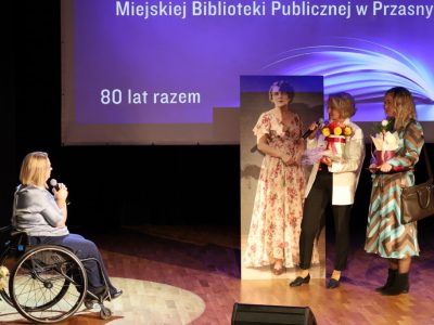 Kobieta w białym żakiecie przemawia do mikrofonu, a stojąca obok niej kobieta w wzorzystej sukience trzyma bukiet żółtych róż i upominek, zwracając się do dyrektor biblioteki