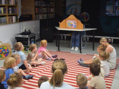 Grupa małych dzieci wraz z opiekunami siedzi na pasiastym, czerwono-białym dywanie w sali bibliotecznej. Wszyscy z zaciekawieniem oglądają teatrzyk obrazkowy Kamishibai, prezentowany w drewnianej skrzynce na stoliku przed nimi. W tle po lewej stronie stoją regały z książkami