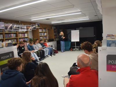 Szerokie ujęcie sali bibliotecznej podczas warsztatów. Joanna Jagiełło stoi w głębi pomieszczenia przy flipcharcie, prowadząc zajęcia dla dużej grupy młodzieży. Uczestnicy siedzą w rzędach wzdłuż regałów z książkami, wypełniając otrzymane materiały szkoleniowe. Na pierwszym planie widać plecy osób siedzących najbliżej obiektywu
