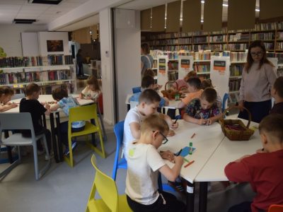 Widok na salę biblioteczną, w której dzieci pracują przy trzech różnych, wielokątnych stołach. Każda grupa wykonuje zadania plastyczne za pomocą kolorowych pisaków i kredek. Między stołami stoi kobieta w okularach, koordynująca przebieg zajęć. W tle widać długie rzędy regałów bibliotecznych oraz wiszące pod sufitem tabliczki informacyjne