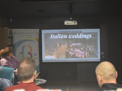 Uczestnicy kawiarenki językowej oglądają prezentację wyświetlaną na ekranie projektora. Na slajdzie widnieje nagłówek „Italian weddings” oraz zdjęcie plenerowego przyjęcia