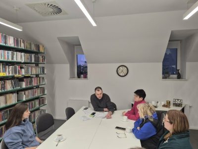Uczestnicy spotkania w bibliotece na poddaszu, sfotografowani pod innym kątem. Mężczyzna siedzący na środku przegląda leżące przed nim dokumenty, gestykulując dłonią. Cztery kobiety siedzące wokół stołu słuchają go. Po prawej stronie widać regał pełen książek, a w głębi stolik z czajnikiem elektrycznym
