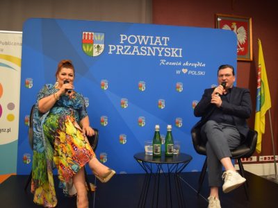 Katarzyna Skrzynecka i prowadzący spotkanie siedzą w czarnych fotelach na scenie. Oboje trzymają mikrofony i uśmiechają się, prowadząc dialog