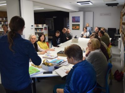 Widok zza pleców prowadzącego w niebieskiej koszuli na uczestników siedzących wzdłuż długiego, podłużnego stołu w bibliotece. Prowadzący prezentuje zebranym materiały plastyczne. Na stole, przed siedzącymi blisko osobami, leżą gotowe, kolorowe rysunki o tematyce jesiennej, taśmy i nożyczki