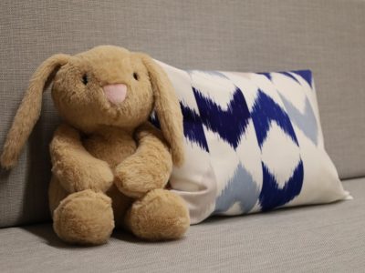 Zbliżenie na beżową, maskotkę obciążeniową w kształcie królika, siedzącą na szarej kanapie obok biało-niebieskiej poduszki we wzory zygzakowate