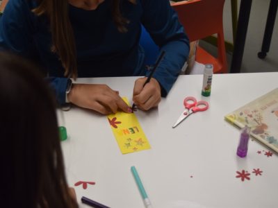 Zbliżenie na dłonie dziecka, które na białym stole ozdabia żółtą papierową zakładkę z kolorowym napisem „LENA”. Obok leżą nożyczki, klej w sztyfcie i małe, wycięte z czerwonego papieru kwiatuszki