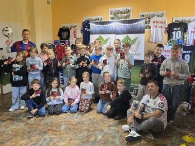 Grupa uśmiechniętych dzieci w sali ozdobionej koszulkami piłkarskimi pozuje do zdjęcia. Uczestnicy trzymają w rękach pamiątkowe karty, a po bokach stoją dwaj mężczyźni w koszulkach klubów piłkarskich – jeden podrzuca piłkę