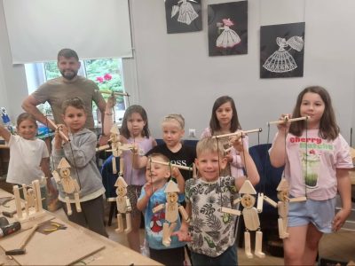 Pamiątkowe zdjęcie z warsztatów stolarskich. Siedmioro uśmiechniętych dzieci trzyma w rękach ukończone drewniane marionetki na sznurkach. Z tyłu stoi brodaty instruktor. W tle widać pracownię