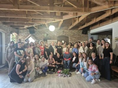Zdjęcie grupowe kilkudziesięciu osób, głównie kobiet, stojących i kucających w przestronnej sali o rustykalnym wystroju. W tle widać ściany z jasnej cegły oraz odsłoniętą, drewnianą konstrukcję dachu. Uczestnicy są uśmiechnięci, ubrani w stroje codzienne. W centralnym punkcie, przed grupą, na podłodze leży bukiet kwiatów