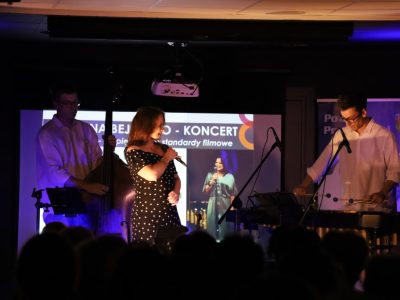 Koncert zespołu Joanna Bejm Trio widziany z perspektywy publiczności, której zaciemnione sylwetki znajdują się na pierwszym planie. Na oświetlonej scenie występuje wokalistka w ciemnej sukience w grochy, śpiewając do mikrofonu. Po jej lewej stronie mężczyzna gra na kontrabasie, a po prawej drugi muzyk gra na wibrafonie. W tle wyświetlany jest napis zapowiadający najpiękniejsze standardy filmowe