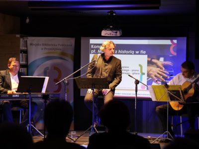 Trzech muzyków występuje razem na scenie podczas koncertu: po lewej stronie mężczyzna gra na instrumentach klawiszowych, na środku wokalista śpiewa do mikrofonu, a po prawej stronie mężczyzna gra na gitarze klasycznej. W tle znajduje się ekran z napisem „Koncert pt. Muzyka z historią w tle”
