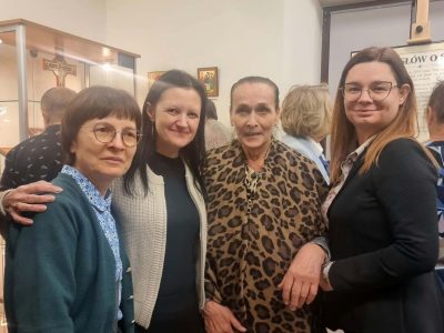 Cztery kobiety uśmiechają się do zdjęcia podczas wydarzenia w galerii. W tle widoczne są szklane gabloty z eksponatami oraz tablica informacyjna na sztaludze