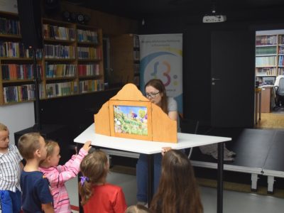 Kobieta w okularach prezentuje ilustrację w drewnianej skrzynce teatrzyku Kamishibai, przed którą stoi i przygląda się grupa małych dzieci. W tle widać regały z książkami oraz wejście do sąsiedniego pomieszczenia biblioteki
