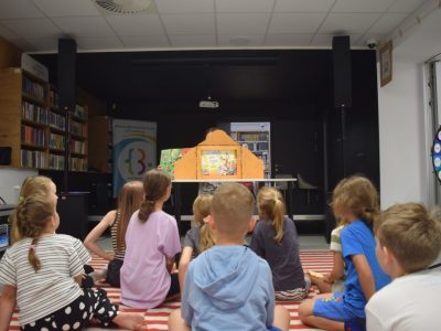 Dzieci siedzą tyłem na pasiastym dywanie, oglądając przedstawienie prezentowane w drewnianej skrzynce teatrzyku Kamishibai, która stoi na stole. W tle widać regał z książkami i kolorowe koło fortuny