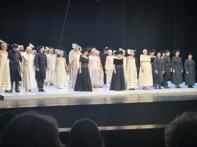 Grupa aktorów w kostiumach z epoki stoi w rzędzie na podwyższeniu sceny podczas spektaklu teatralnego. Część obsady ma na sobie jasne, długie suknie i charakterystyczne płaskie nakrycia głowy, pozostali ubrani są w ciemne, historyczne płaszcze i habity. Scena jest zaciemniona, a z przodu widoczne są zarysy głów publiczności