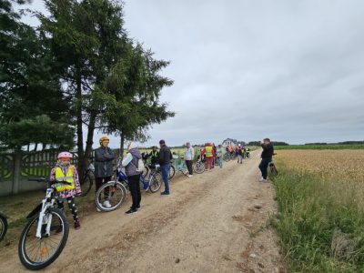Rowerzyści uczestniczący w rajdzie zatrzymali się na odpoczynek na poboczu piaszczystej drogi polnej. Po prawej stronie rozciąga się duże pole dojrzałego zboża, a po lewej rosną gęste iglaki za betonowym płotem. Uczestnicy stoją przy swoich rowerach i rozmawiają