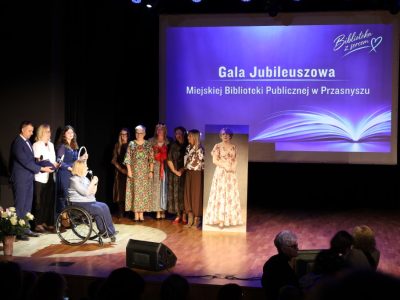 Szerokie ujęcie na scenę, gdzie obok dyrektor biblioteki na wózku inwalidzkim oraz prowadzących wydarzenie stoi grupa pięciu kobiet w eleganckich sukienkach. Obok nich umieszczono stojący portret kobiety w retro sukni, a w tle wyświetla się ekran z napisem „Gala Jubileuszowa Miejskiej Biblioteki Publicznej w Przasnyszu”