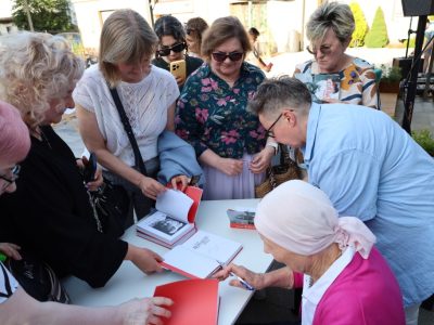 Anna Nehrebecka siedzi przy białym stoliku i podpisuje książki dla zgromadzonych wokół niej czytelniczek. Kobiety z zaciekawieniem przyglądają się autorce, trzymając w dłoniach egzemplarze książek oraz telefony. Na stole leżą otwarte publikacje przygotowane do podpisu