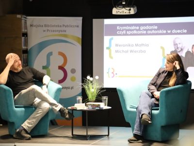 Weronika Mathia i Michał Wierzba podczas rozmowy na scenie. Siedzą naprzeciwko siebie w nowoczesnych, turkusowych fotelach. Między nimi stoi mały stolik z książkami i kwiatami. W tle znajduje się ekran z prezentacją przedstawiającą zdjęcia i nazwiska autorów oraz logotyp Miejskiej Biblioteki Publicznej w Przasnyszu
