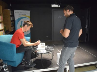 Daniel Kocuj siedzi w turkusowym fotelu na niskim podwyższeniu i podpisuje egzemplarz książki leżący na okrągłym, czarnym stoliku. Autor ma na sobie czerwoną koszulkę, ciemne spodnie i sportowe buty. Przed podwyższeniem stoi odwrócony tyłem mężczyzna w ciemnej koszulce polo i szarych spodniach, który czeka na autograf. W tle widoczny jest baner z logo Miejskiej Biblioteki Publicznej w Przasnyszu