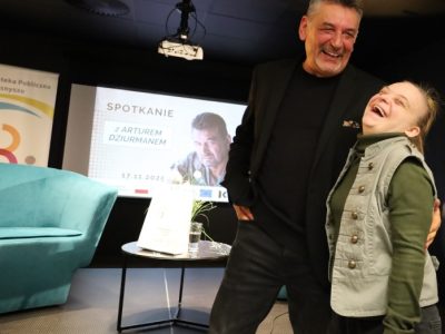 Radosny kadr ze spotkania. Artur Dziurman i stojąca obok niego kobieta w zielonej kamizelce śmieją się szeroko, patrząc w stronę obiektywu. W tle widoczna jest scena z fotelem, stolikiem oraz ekranem informacyjnym