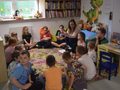 Dzieci siedzą w kręgu na wzorzystym, kolorowym dywanie w kąciku dziecięcym w bibliotece. Przed nimi kuca kobieta w okularach, która trzyma w dłoniach małą kartkę i pokazuje ją grupie. W tle widać niskie regały z książkami, pluszowe zabawki na parapecie okna oraz dużą naklejkę z zielonym smokiem na ścianie