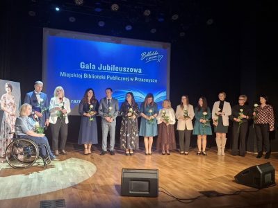 Szerokie ujęcie sceny, na której stoi kilkanaście osób trzymających białe róże. Po lewej stronie na wózku siedzi dyrektor biblioteki przemawiająca do mikrofonu. W tle wyświetla się slajd „Gala Jubileuszowa Miejskiej Biblioteki Publicznej w Przasnyszu”