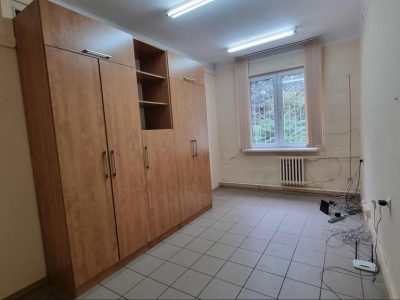 Puste, wąskie pomieszczenie z kafelkami na podłodze i jasnymi ścianami. Po lewej stronie stoi duża, wysoka szafa z jasnego drewna z wbudowanymi regałami. Na wprost znajduje się okno z żaluzjami pionowymi, pod którym zamontowany jest żeliwny kaloryfer. W prawym rogu na podłodze leży splątana wiązka kabli sieciowych oraz router